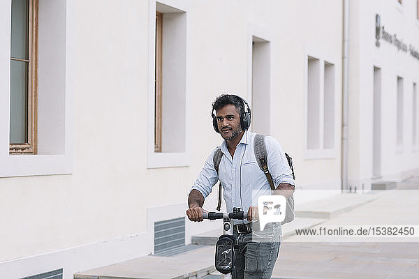 Casual businessman wearing headphones riding e-scooter in the city