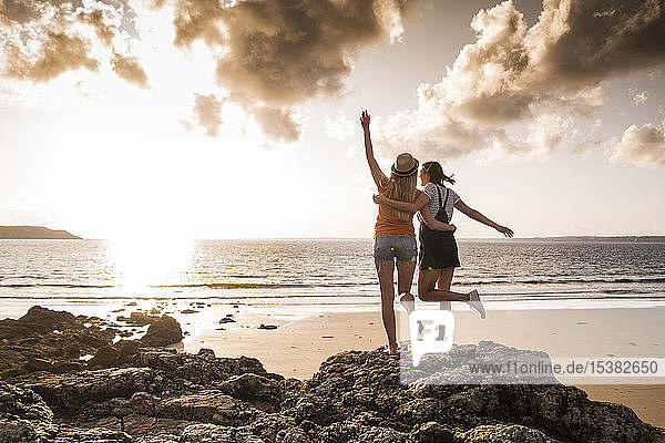 Two girlfriends standing on rocky beach  waving at sunset  rear view