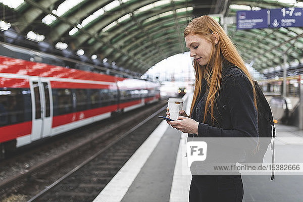 Rothaarige junge Frau mit Kaffee zum Warten am Bahnsteig mit Smartphone  Berlin  Deutschland