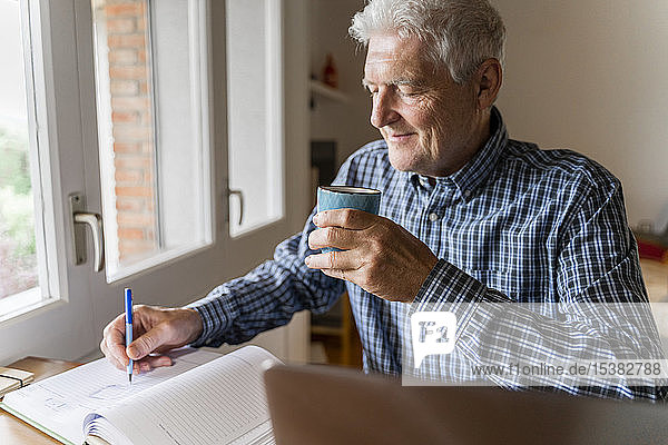 Älterer Mann sitzt zu Hause vor dem Laptop und macht sich Notizen
