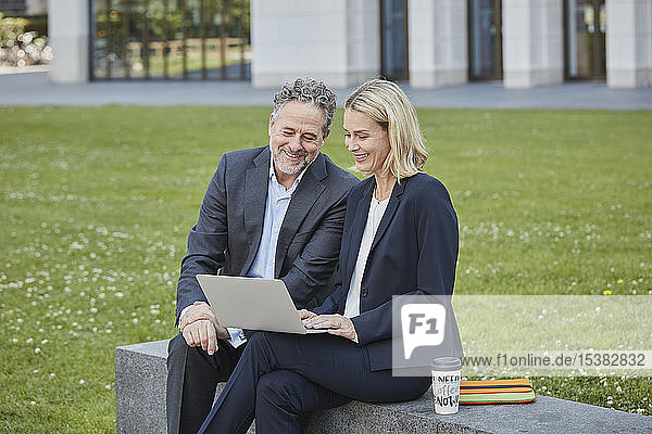 Businesswoman and businessman sitting on a wall in the city using laptop
