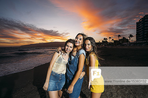 Porträt von drei glücklichen Freundinnen am Strand bei Sonnenuntergang