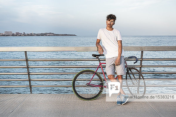 Junger Mann mit Fahrrad  auf der Brücke am Meer stehend