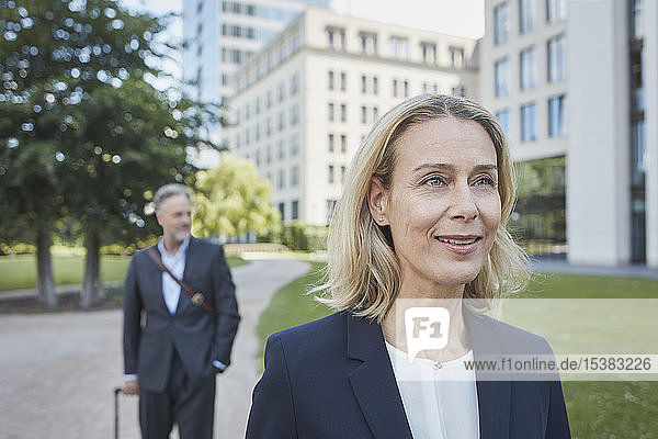 Porträt einer lächelnden blonden Geschäftsfrau in der Stadt mit einem Geschäftsmann im Hintergrund