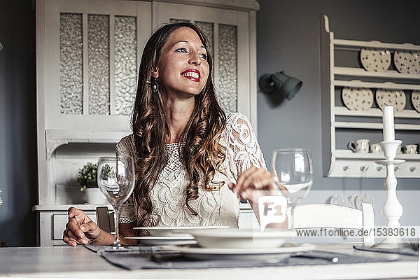 Porträt einer lächelnden jungen Frau  die am gedeckten Tisch in einer Küche im Landhausstil sitzt