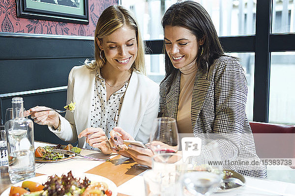 Zwei glückliche Geschäftsfrauen in einem Restaurant  die sich gemeinsam ein Smartphone anschauen