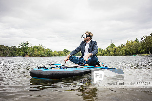 Businessman wearing VR glasses sitting on SUP board on a lake