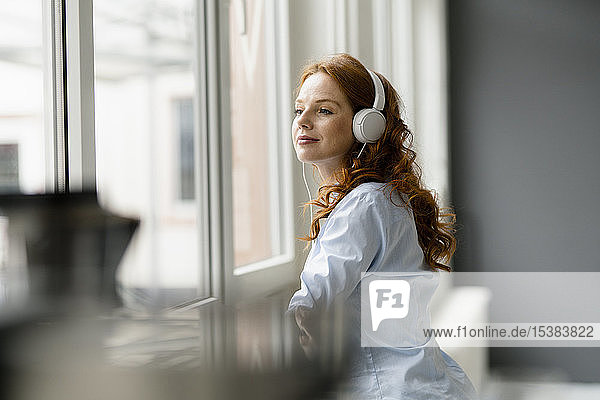 Porträt einer rothaarigen Geschäftsfrau  die mit Kopfhörern Musik hört und dabei aus dem Fenster schaut