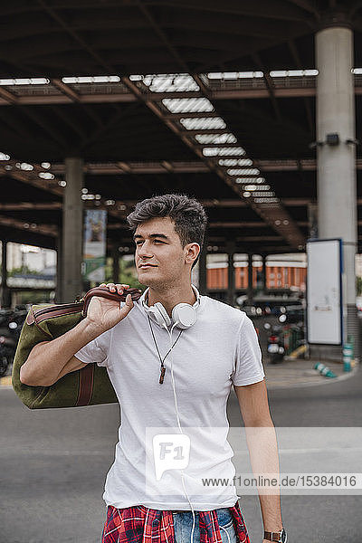 Junger Mann mit einer Tasche am Bahnhof mit Kopfhörern