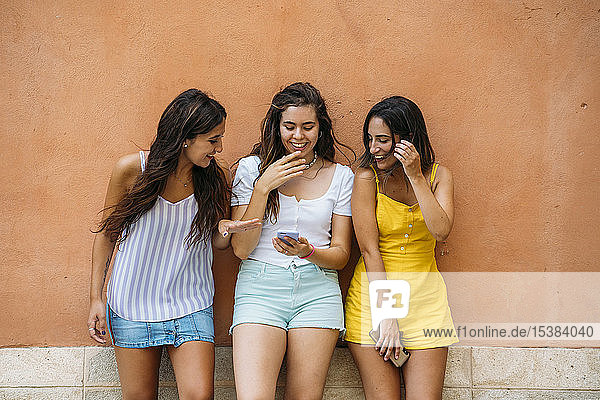 Three happy female friends standing at a wall sharing smartphone