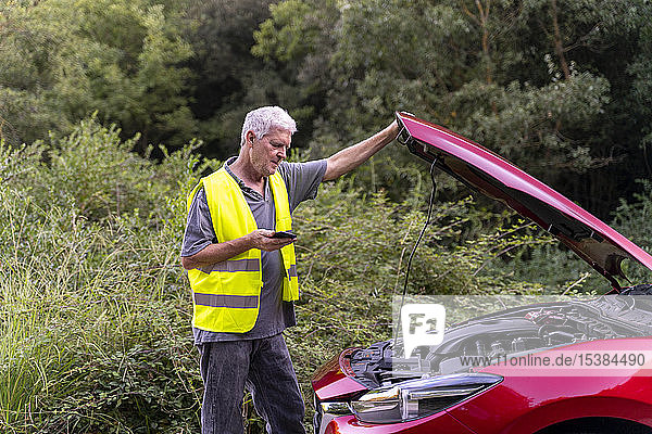 Senior man standing at his broken car wearing a safety vest and using his smartphone