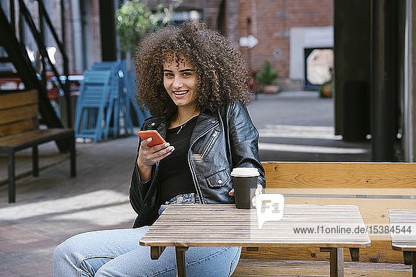 Porträt einer lächelnden Teenagerin  die auf einer Bank sitzt  mit Kaffee zum Mitnehmen und Handy