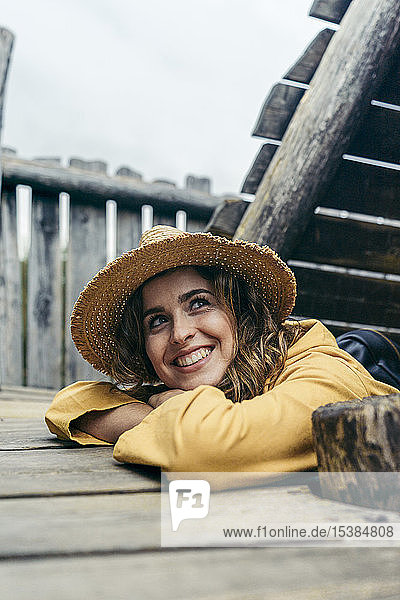 Junge lächelnde Frau mit gelbem Mantel und Hut auf einer Holztreppe  seitwärts blickend