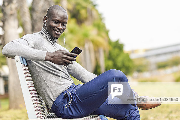 Porträt eines lächelnden Geschäftsmannes  der auf einer Bank sitzt und mit Kopfhörern und Smartphone Musik hört
