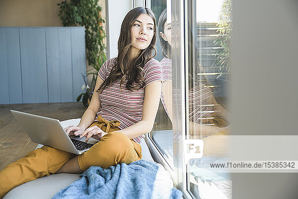 Junge Frau sitzt zu Hause mit Laptop am Fenster