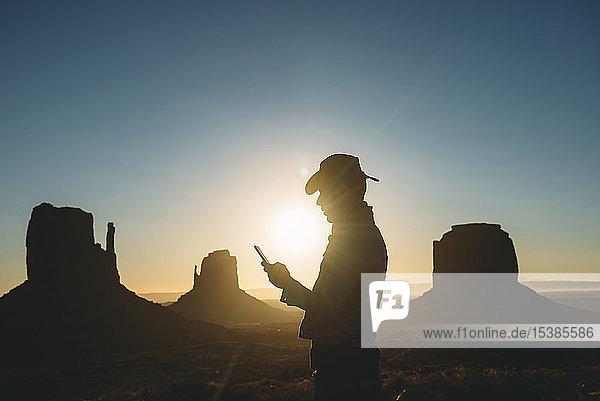USA  Utah  Monument Valley  Silhouette eines Mannes mit Cowboyhut  der bei Sonnenaufgang auf sein Handy schaut