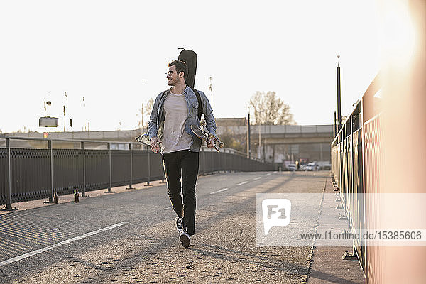 Lächelnder junger Mann mit Gitarrenkoffer und Skateboard geht auf einer Brücke in der Stadt