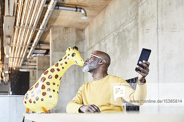 Reifer Geschäftsmann sitzt am Schreibtisch im Büro mit einem Handy  das eine Giraffenfigur küsst
