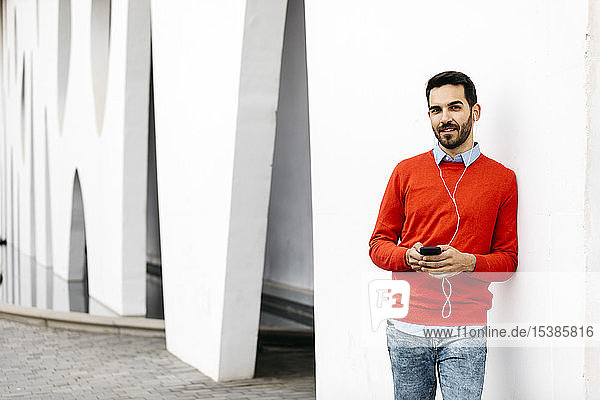 Lässiger Geschäftsmann  der sich an die Wand lehnt und Kopfhörer und Smartphone benutzt