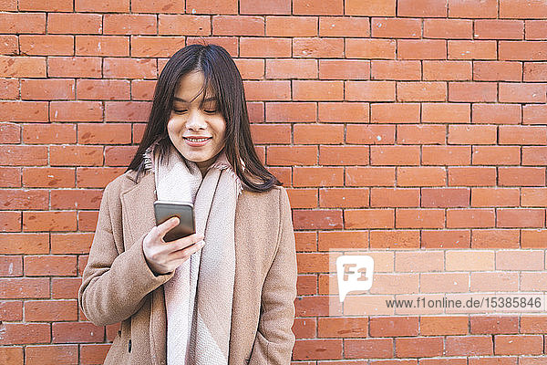 Lächelnde junge Frau benutzt Handy an Ziegelmauer