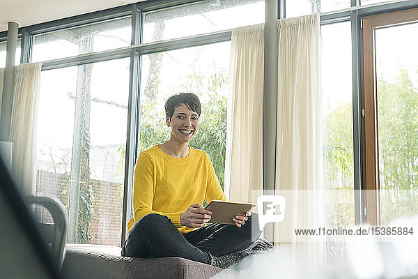 Porträt einer glücklichen Frau mit digitalem Tablett  die auf der Couch im Wohnzimmer sitzt