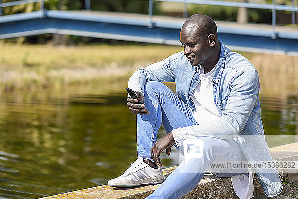Mann in lässiger Denim-Kleidung mit Smartphone am Fluss sitzend