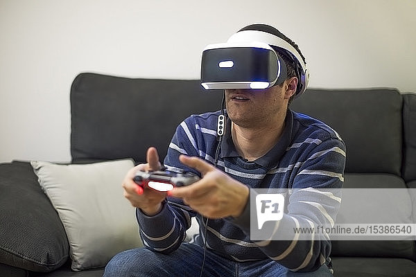 Junger Mann mit VR-Brille sitzt zu Hause auf der Couch und spielt Videospiel