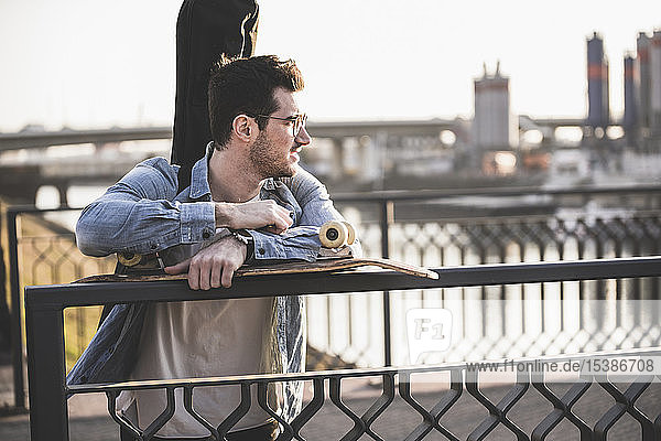 Young man with guitar case and skateboard on a bridge in the city looking around