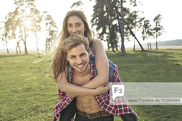 Porträt eines glücklichen jungen Mannes  der seine Freundin huckepack auf der Wiese trägt