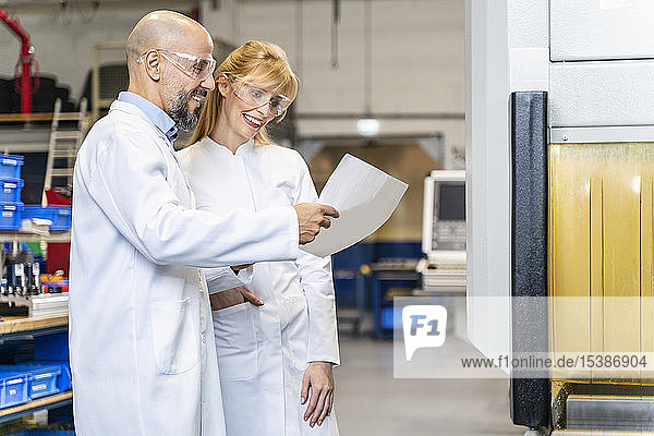 Zwei glückliche Techniker mit Laborkittel und Schutzbrille sehen sich den Plan in der Fabrik an