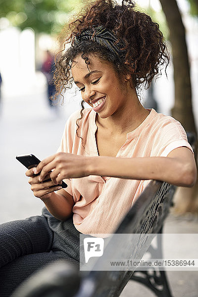 Lächelnde junge Frau sitzt auf einer Bank und benutzt ihr Handy