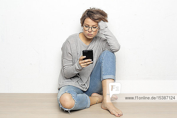 Porträt einer jungen Frau mit drahtlosen Kopfhörern  die auf dem Boden sitzt und auf ihr Handy schaut