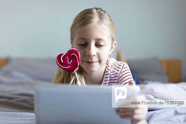 Porträt eines blonden  auf dem Bett liegenden Mädchens mit Lolli mit digitalem Tablett