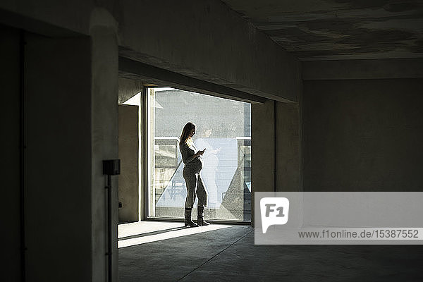 Schwangere Geschäftsfrau steht am Bürofenster und benutzt ein Smartphone