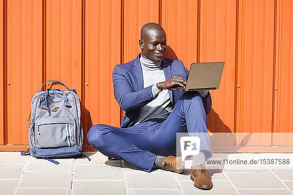 Modischer Geschäftsmann sitzt mit Laptop vor oranger Wand