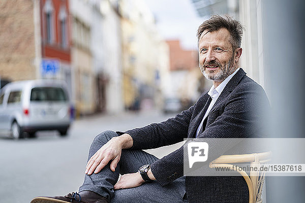 Portrait of content mature businessman with greying beard sitting on chair in the city