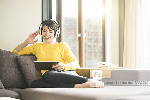 Portrait of happy woman sitting on the couch at home listening music with headphones while looking at tablet