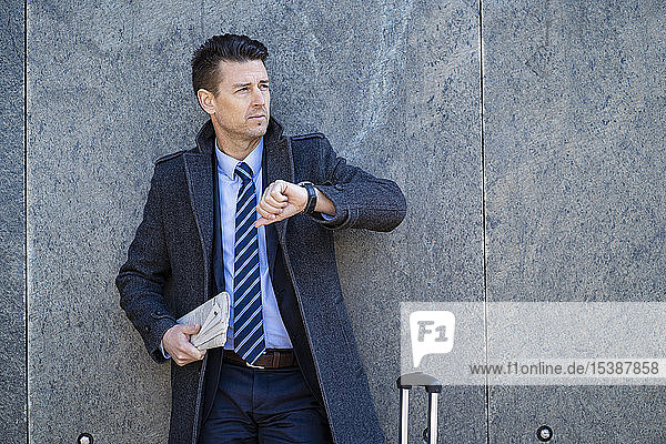 Businessman with suitcase standing at a wall checking the time