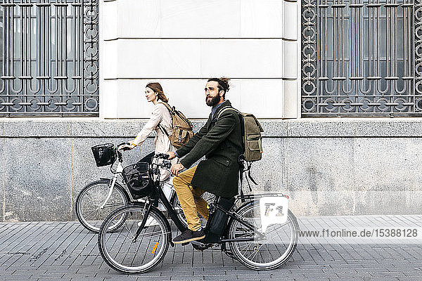 Ein Paar fährt mit E-Bikes in der Stadt an einem Gebäude vorbei