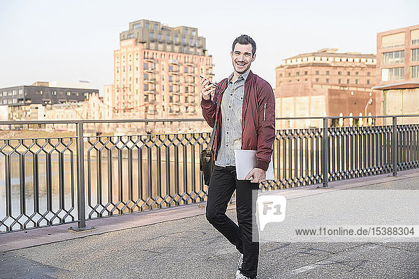 Glücklicher junger Mann mit Tablette  Handy und Tasche auf einer Brücke in der Stadt