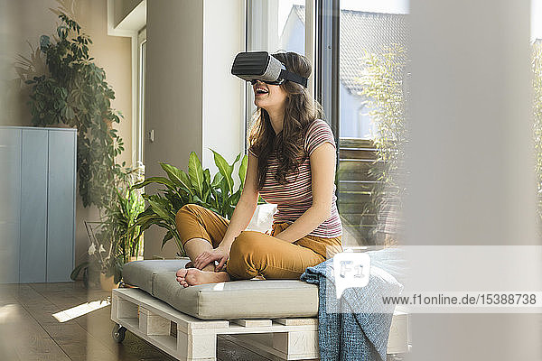 Glückliche junge Frau  die zu Hause am Fenster sitzt und eine VR-Brille trägt