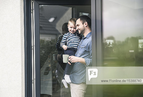 Father with cup of coffee carrying son at terrace door at home