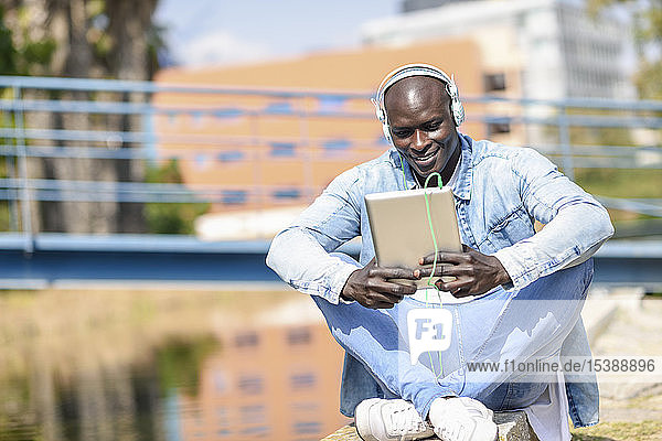 Mann in lässiger Jeansbekleidung  der im Freien mit Kopfhörern und digitalem Tablet Musik hört