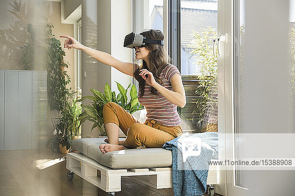 Junge Frau  die zu Hause am Fenster sitzt und eine VR-Brille trägt