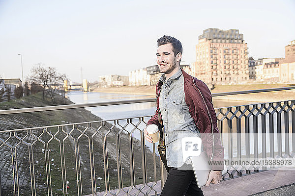 Smiling young man with tablet  bag and takeaway coffee on a bridge in the city