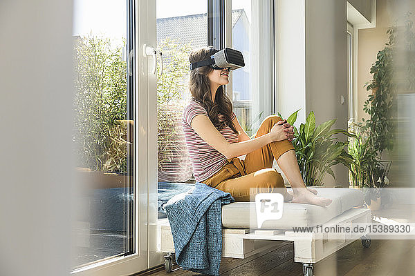 Young woman sitting at the window at home wearing VR glasses