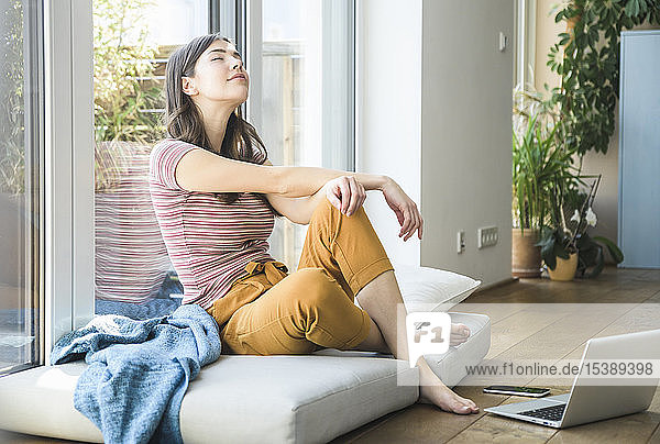 Relaxed young woman sitting at the window at home with laptop