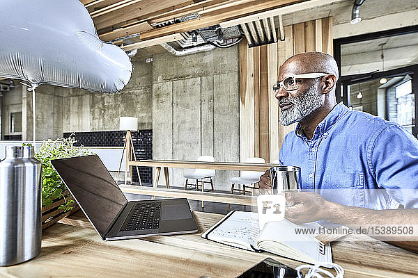 Reifer Geschäftsmann sitzt am Tisch im modernen Büro mit Wolkenballon und Laptop