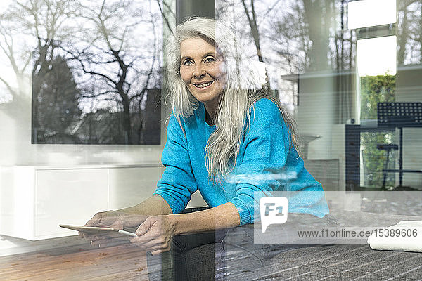 Porträt einer lächelnden reifen Frau mit digitalem Tablett  die zu Hause auf der Couch sitzt und aus dem Fenster schaut
