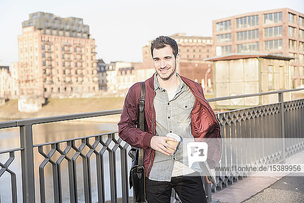Smiling young man with tablet  bag and takeaway coffee on a bridge in the city